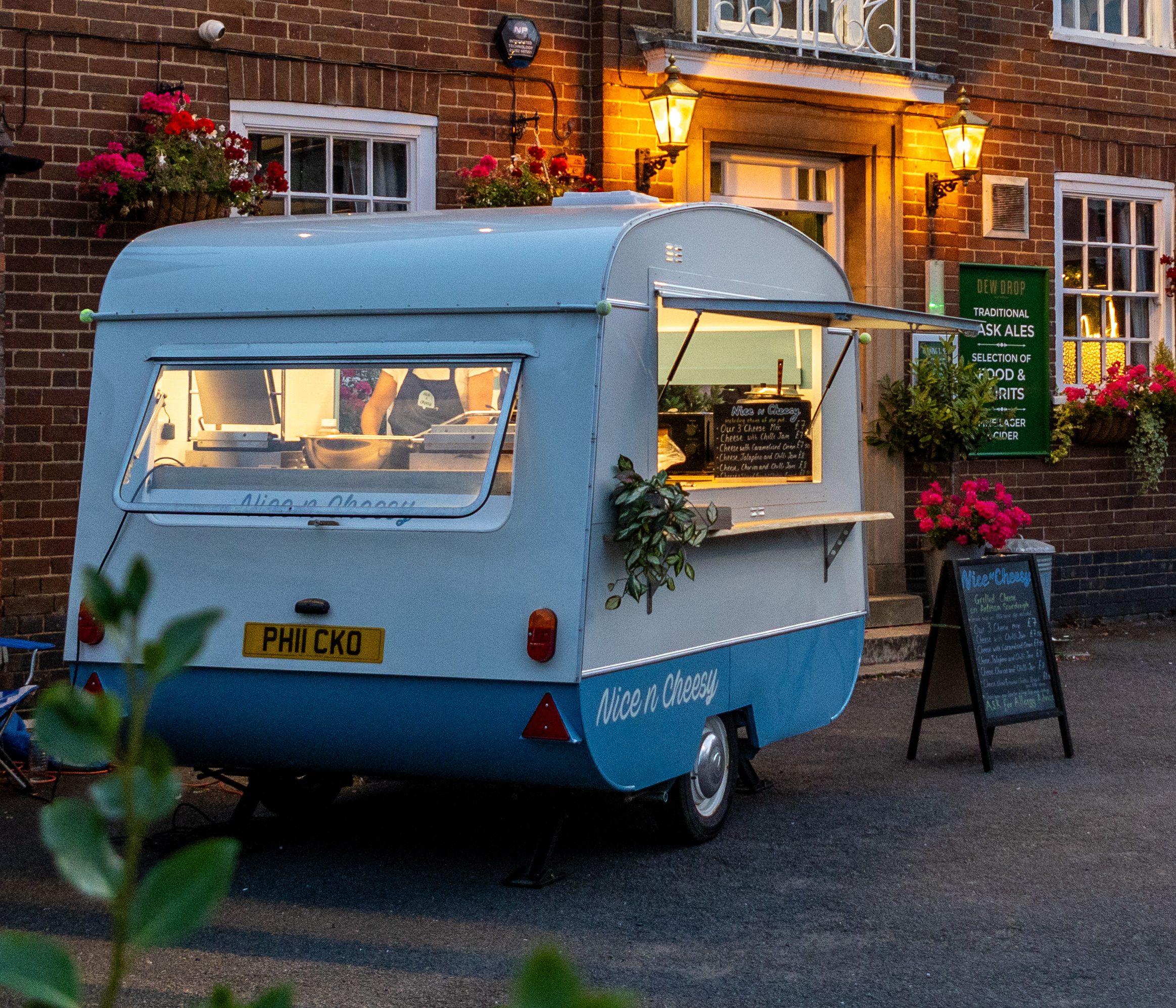 A vintage food caravan named 'Nice n Cheesy' parked outside a brick building, featuring a warm glow from the interior lights and surrounded by flower baskets, ready to serve artisan toasties for a corporate event.