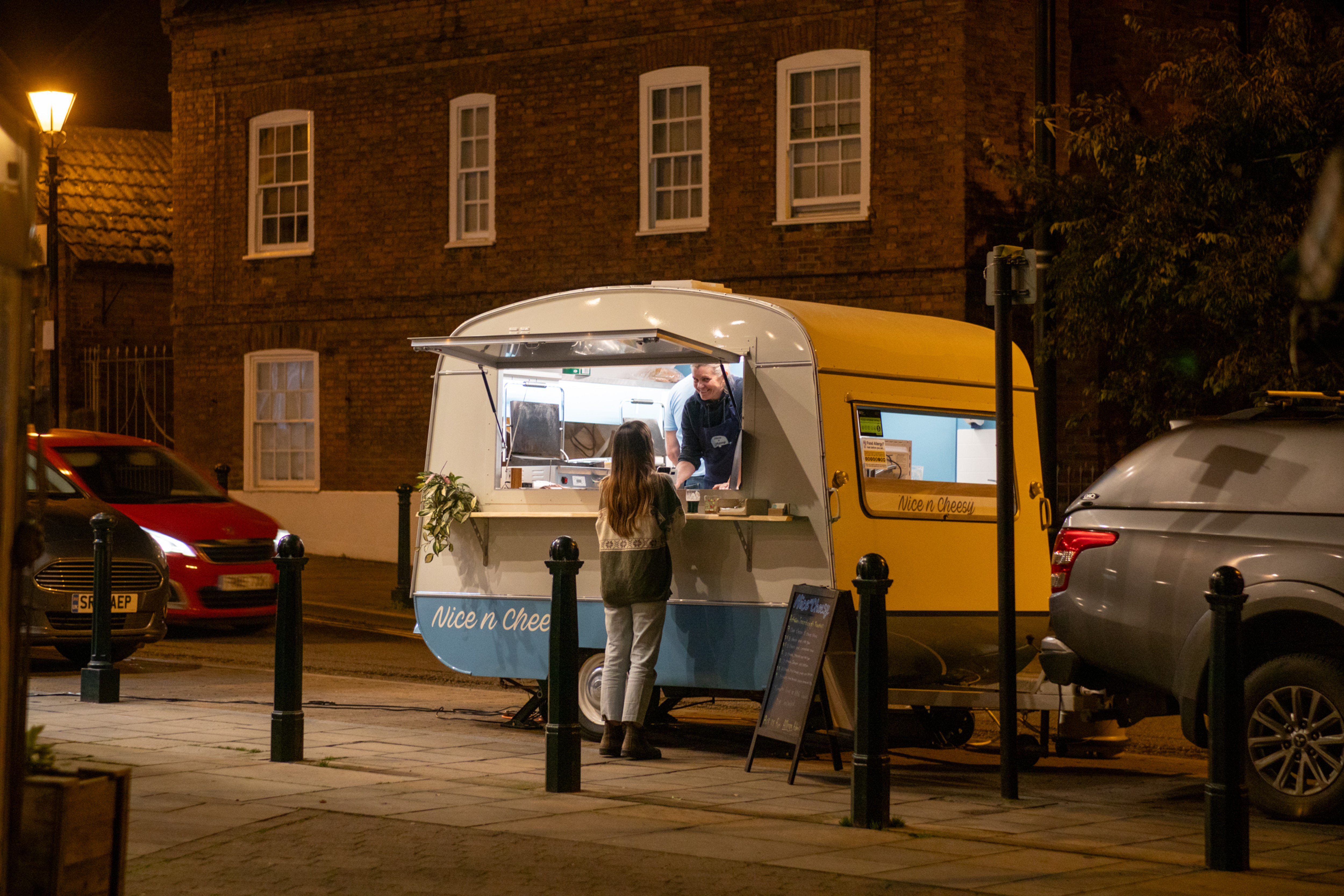A vintage food caravan named 'Nice n Cheesy' serving artisan toasties at night, with a happy customer interacting with the vendor.